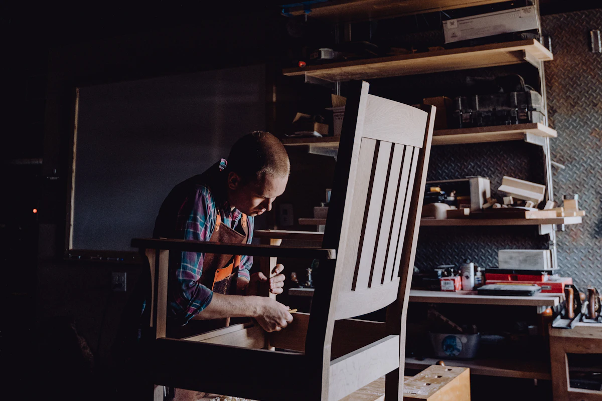 A maker in a workshop assembling a chair, tools racked on the wall behind
